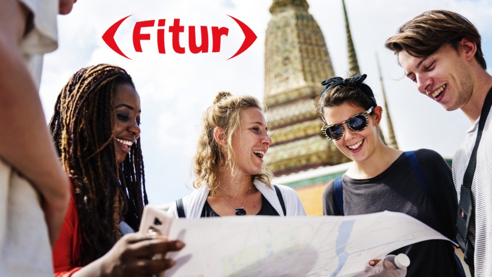 cinco jóvenes con un mapa y el logo de FITUR, monumento de fondo
